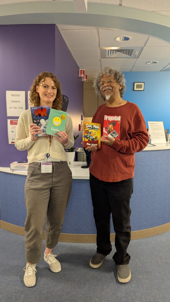 A white woman & a Black man pose in front of a hospital check-in desk. They are smiling & each is holding two young adult graphic novels (she is holding a volume of BONE & a copy of SMILE; he is holding a volume of DOG MAN & a volume of DRAGON BALL).